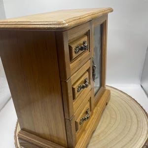 May include: A wooden jewelry box with three drawers and a glass-fronted door. The box is made of light brown wood with ornate metal drawer pulls. The jewelry box is displayed on a wooden slice.