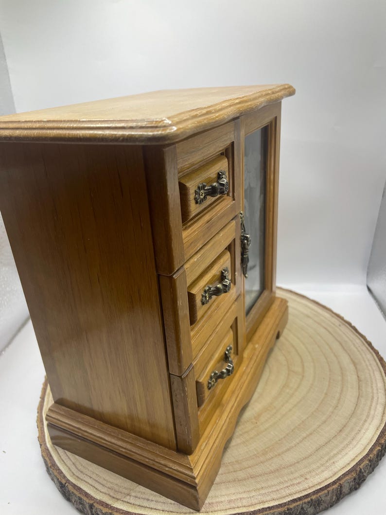 May include: A wooden jewelry box with three drawers and a glass-fronted door. The box is made of light brown wood with ornate metal drawer pulls. The jewelry box is displayed on a wooden slice.