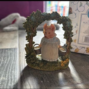 May include: A Peter Rabbit arch figurine from The World of Beatrix Potter. The figurine features a pig character wearing a dress and apron, holding two buckets. The arch is decorated with green foliage and small yellow flowers.