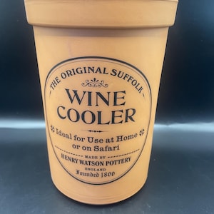 May include: A terracotta-colored wine cooler with a black circular label. The label reads "THE ORIGINAL SUFFOLK WINE COOLER" and "HENRY WATSON POTTERY ENGLAND Founded 1800". The cooler is cylindrical and has a slightly flared rim.