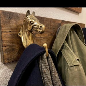 以下が含まれることがあります： 真鍮製の馬の頭のフックが付いた素朴な木製のコートラック。 ネイビーブルーのコート、柄のスカーフ、オリーブグリーンのジャケットが掛かっています。 馬の頭のフックは、ユニークで乗馬にインスパイアされたタッチを追加します。