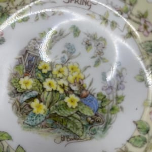 Puede incluir: Un plato decorativo con temática primaveral. El plato presenta una ilustración detallada de un ratón con una chaqueta azul entre flores amarillas y follaje verde. El plato tiene un borde color crema con detalles florales y las palabras "Brambly Hedge SPRING".