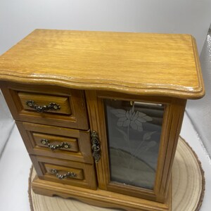 May include: A wooden jewelry box with three small drawers on the left and a glass door on the right. The glass door has a floral design etched into it. The box is made of light brown wood and sits on a wooden base.