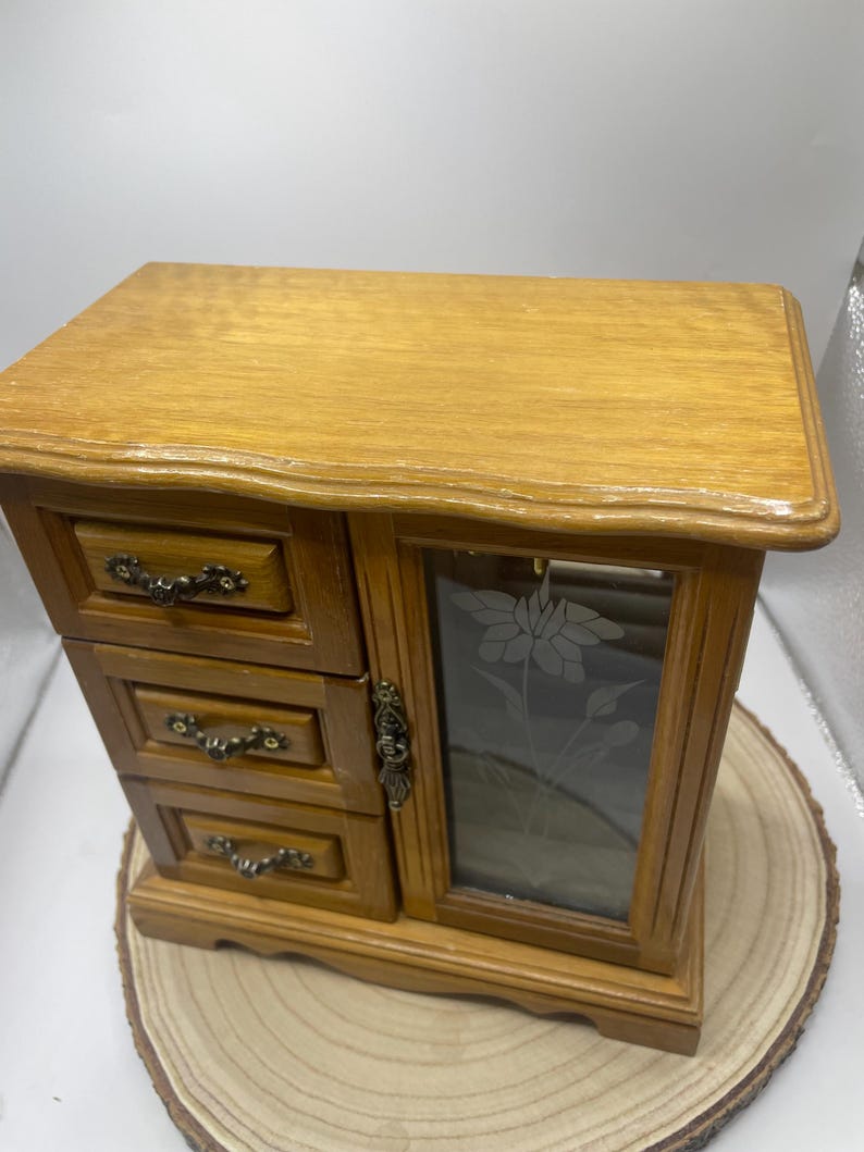 May include: A wooden jewelry box with three small drawers on the left and a glass door on the right. The glass door has a floral design etched into it. The box is made of light brown wood and sits on a wooden base.