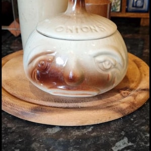 May include: A ceramic onion-shaped container with a lid, featuring a face design with brown accents. The word "ONIONS" is on the lid. It sits on a wooden plate. Other ceramic items are in the background.