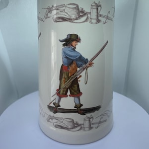Könnte beinhalten: Ein weißer Bierkrug aus Keramik mit einer detaillierten Illustration einer historischen Figur in historischer Kleidung, die ein langes Gewehr hält. Das Design umfasst Hüte, Schwerter und Krüge. Der Krug ist zylindrisch.