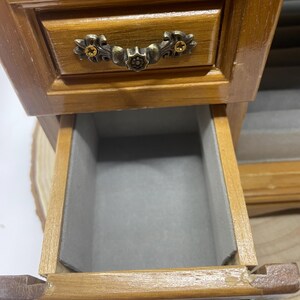 May include: A small, wooden jewelry box with a drawer pulled open. The box is stained a warm brown color and features ornate metal hardware. The drawer interior is lined with gray fabric, providing a soft space for storing jewelry.