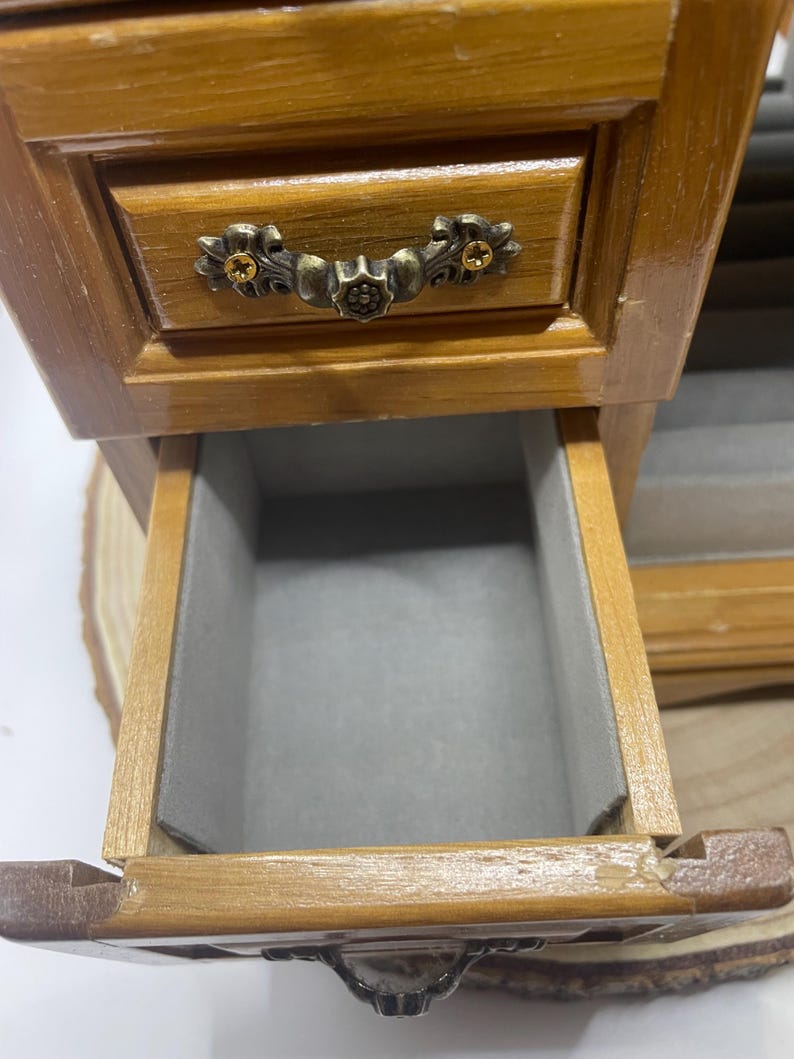 May include: A small, wooden jewelry box with a drawer pulled open. The box is stained a warm brown color and features ornate metal hardware. The drawer interior is lined with gray fabric, providing a soft space for storing jewelry.