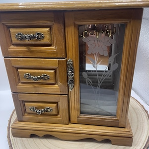May include: A wooden jewelry box with three drawers and a mirrored door. The door features a frosted floral design. The box has ornate drawer pulls and a decorative latch. The box is made of light brown wood and sits on a wooden base.