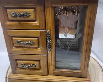 May include: A wooden jewelry box with three drawers and a mirrored door. The door features a frosted floral design. The box has ornate drawer pulls and a decorative latch. The box is made of light brown wood and sits on a wooden base.
