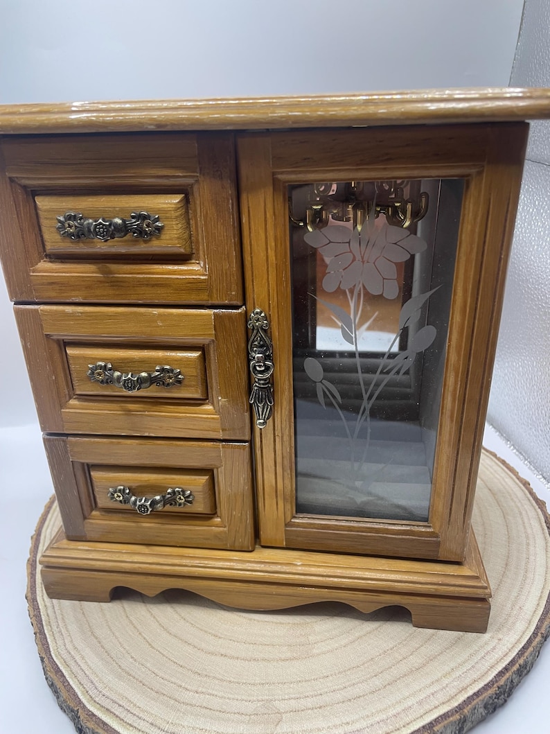 May include: A wooden jewelry box with three drawers and a mirrored door. The door features a frosted floral design. The box has ornate drawer pulls and a decorative latch. The box is made of light brown wood and sits on a wooden base.