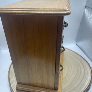 May include: A small wooden jewelry box with three drawers. The box is made of light brown wood with a darker brown base and top. The drawers have ornate metal handles. The box is sitting on a wooden slice.