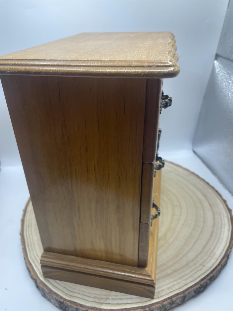 May include: A small wooden jewelry box with three drawers. The box is made of light brown wood with a darker brown base and top. The drawers have ornate metal handles. The box is sitting on a wooden slice.