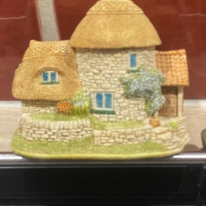 Könnte beinhalten: Miniatur-Figur eines Cottage mit Strohdach, Steinmauern und kleinen blauen Fenstern. Die Figur hat einen Schornstein und einen kleinen Garten. Die Farben sind in gedämpften Beige-, Grün- und Brauntönen gehalten.