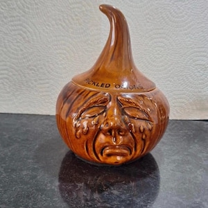 May include: A brown ceramic jar shaped like an onion with a face, labeled "PICKLED ONIONS." The jar has a lid and a long, curved top. The face has closed eyes and a sad expression.