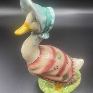Puede incluir: Figura de pato de cerámica con un gorro azul claro y un vestido a rayas rosas y blancas. El pato tiene un pico amarillo y está sobre una base verde. La figura es un objeto decorativo.