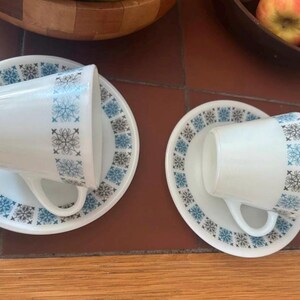 May include: Two white teacups and saucers with a snowflake design in blue and black. The cups are resting on their saucers, set against a brown tile surface. A wooden bowl and apples are in the background.