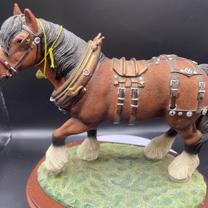May include: A detailed figurine of a brown horse with a black mane and tail, wearing a harness. The horse has white fur on its legs and stands on a green grassy base with a wooden border. The horse is posed in a walking stance.