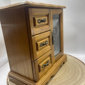May include: A wooden jewelry box with three small drawers and a glass door. The box is a light brown color with ornate metal drawer pulls. The glass door has a floral design etched into it. The box is sitting on a wooden slice.