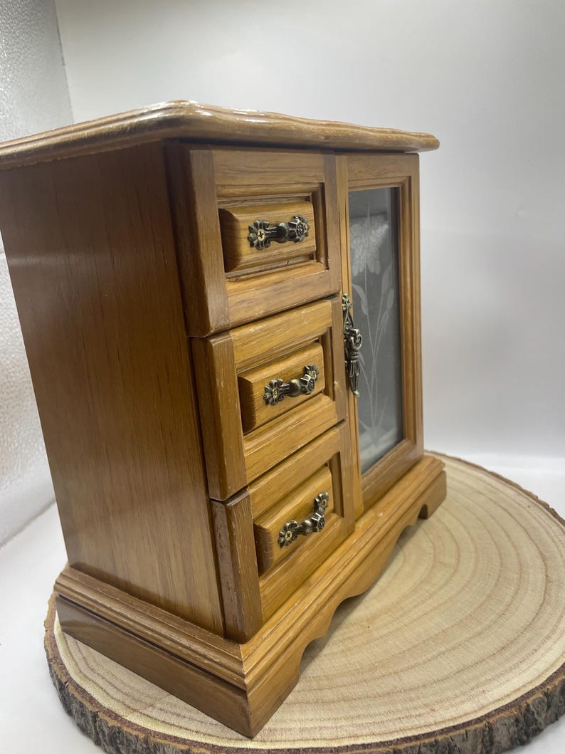May include: A wooden jewelry box with three small drawers and a glass door. The box is a light brown color with ornate metal drawer pulls. The glass door has a floral design etched into it. The box is sitting on a wooden slice.