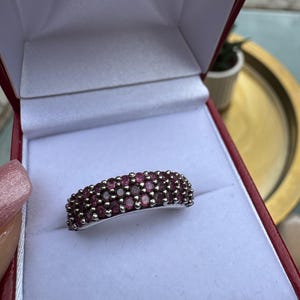 Puede incluir: Un anillo de banda en tono plateado con tres filas de pequeñas piedras preciosas redondas de color rosa intenso dispuestas en racimo. El anillo se muestra dentro de un joyero rojo con forro interior blanco, destacando el diseño de varias piedras.
