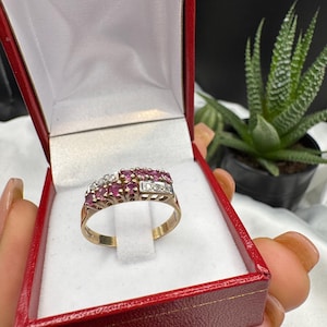 Könnte beinhalten: Ein goldener Ring mit kleinen, runden, roten und klaren Edelsteinen, präsentiert in einer roten Schmuckschatulle mit weißem Innenraum. Der Ring wird in der Hand einer Person gehalten.