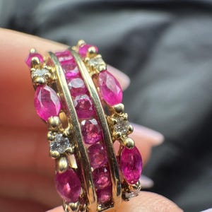 Puede incluir: Un anillo de oro con un canal central engastado con rubíes rosas redondos, flanqueado por dos filas exteriores que contienen una alternancia de rubíes rosas talla marquesa y pequeños diamantes blancos redondos en un diseño de banda.