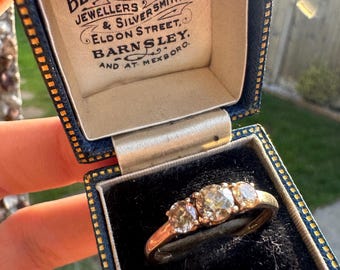 May include: A gold three-stone ring featuring three round-cut diamonds set in a dark blue velvet-lined jewellery box. The box lid interior features printed text reading Ben Harral Ltd, Jewellers & Silversmiths, Eldon Street, Barnsley and at Mexboro.