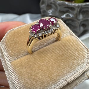 May include: A gold ring with three oval rubies and smaller clear gemstones. The ring is presented on a velvet ring box, highlighting the jewellery's design and colour.