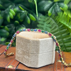 Pode incluir: Uma pulseira de ténis em ouro com um padrão repetitivo de pedras preciosas de corte oval vermelho, azul e verde, intercaladas com pequenas pedras de acento de corte redondo e transparente numa corrente de metal.