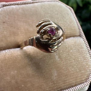 May include: A gold ring featuring two sculpted hands meeting in the centre to hold a small, round, faceted pink gemstone. The band of the ring is smooth and tapers towards the back, with the hands forming the main decorative setting of the piece.