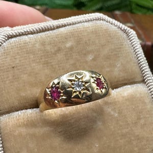 Puede incluir: Un anillo de oro con una piedra preciosa central transparente flanqueada por dos piedras rojas de corte marquesa. Cada piedra está engastada en un grabado en forma de estrella sobre la banda de oro lisa y redondeada, creando un diseño celestial.