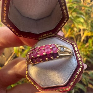 Può includere: Un anello d'oro con una fila di rubini rossi vibranti e un piccolo diamante. L'anello è esposto all'interno di un portagioie bordeaux con finiture dorate, che ne evidenzia l'eleganza e il design. L'anello è un gioiello.