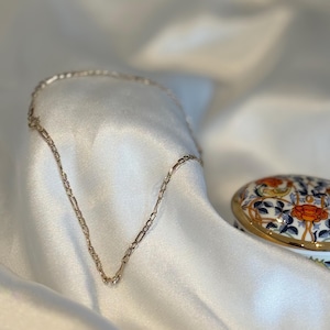 May include: A fine gold-coloured necklace with a linked chain design lies on a white satin surface. A small, decorative trinket box with a floral pattern in orange, blue, and white is in the background. The jewellery is elegant and simple.