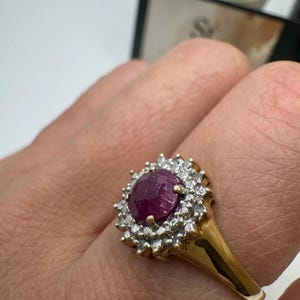May include: A gold ring with a large, round, deep red gemstone encircled by smaller, clear stones. The ring's band is polished gold, and the gemstone setting is detailed. The ring's design is classic and elegant.