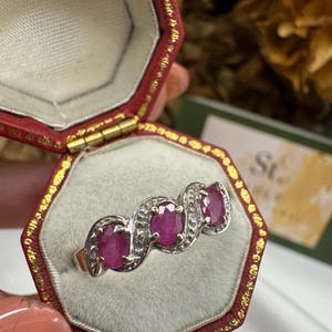 May include: A gold and silver ring with three oval-cut rubies. The rubies are set in a wave-like pattern, surrounded by small, clear stones. The ring is displayed in a red jewelry box with a gold trim.