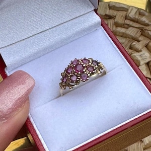 May include: A silver ring featuring a cluster of multiple round, faceted pink and magenta gemstones arranged in a dome shape. The metal band has a polished finish and supports the gemstone setting, which is presented inside a white velvet ring box.