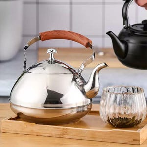 Op de afbeelding: Een glanzende roestvrijstalen theepot met een houten handvat en een klein glazen kopje op een houten dienblad. De theepot heeft een afgeronde body en een gebogen tuit. Een zwarte theepot staat op de achtergrond.