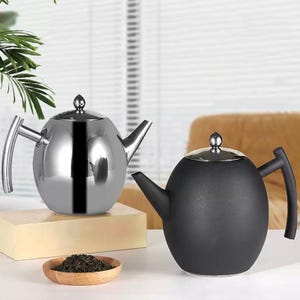 Op de afbeelding: Twee theepotten, een glanzend zilveren en de andere matzwart, worden getoond. Beide hebben een ronde body, een tuit en een handvat. De zilveren theepot staat op een lichtgekleurd blok, met een kleine houten kom met theebladeren ervoor.