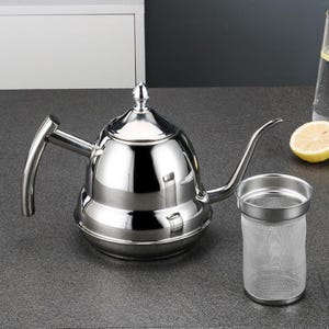 Può includere: Una lucida teiera a collo di cigno in acciaio inossidabile con un beccuccio lungo e curvo e un infusore per tè abbinato. La teiera ha un corpo arrotondato e un manico. Una fetta di limone e un bicchiere d'acqua sono sullo sfondo.