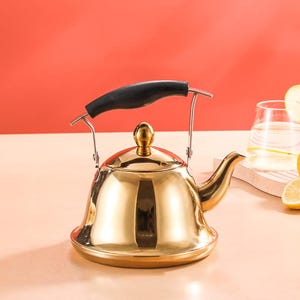 May include: A shiny gold-colored tea kettle with a black handle and a spout. The kettle has a rounded base and a lid with a small knob. The background is a solid red color.