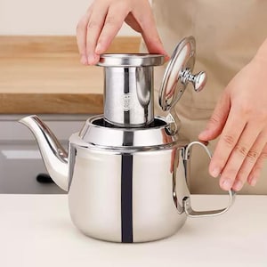 Puede incluir: Una tetera de acero inoxidable pulido con un infusor y tapa extraíbles. La tetera tiene un pico curvo y un asa. El infusor se está colocando dentro de la tetera. La tetera está sobre una superficie blanca.