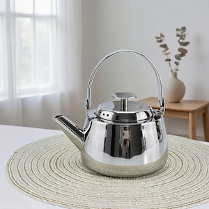 May include: A shiny, stainless steel tea kettle with a curved handle and a spout. The kettle has a silver base and a lid with a small handle. It sits on a round, beige placemat. The background includes a window and a small wooden table.
