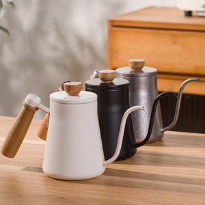 May include: Three gooseneck kettles in white, black, and silver, with wooden handles and lids. The kettles are designed for pour-over coffee brewing, with a long, curved spout for precise pouring. The kettles are displayed on a wooden surface.