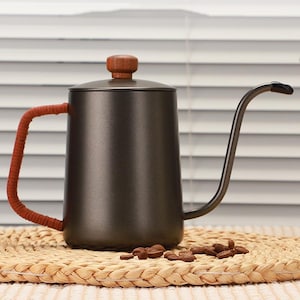 May include: A dark grey gooseneck kettle with a wooden knob and a red wrapped handle. The kettle is designed for pour-over coffee brewing. Coffee beans are scattered on a woven mat.