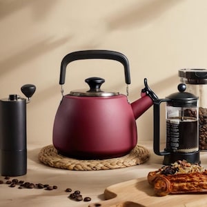 Pode incluir: Uma chaleira bordô com alça e bico pretos está sobre um tapete tecido. Um moedor de café preto e uma prensa francesa também são visíveis, junto com grãos de café e uma tábua de corte de madeira. Tema de café e chá.