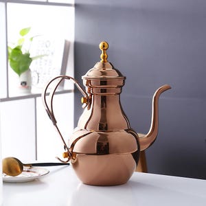 Op de afbeelding: Een koperkleurige, sierlijke theepot met een lange, gebogen tuit en een afgeronde body. De theepot heeft een handvat en een decoratieve knop op het deksel. Een goudkleurige lepel rust op een klein bord. De theepot is waarschijnlijk voor het serveren van thee of koffie.