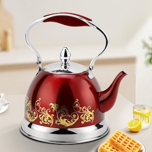Puede incluir: Una tetera roja con asa y base plateadas. La tetera presenta un diseño floral dorado en el cuerpo. El asa tiene un agarre rojo. La tetera está sobre una mesa con un vaso de agua y un plato de gofres.