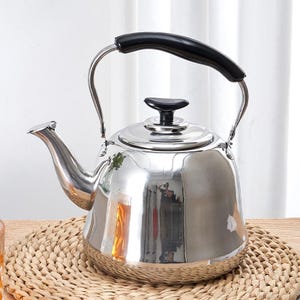 May include: A shiny, stainless steel tea kettle with a black handle and spout. The kettle has a curved handle and a small, black knob on the lid. It sits on a woven mat.