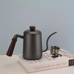 Może przedstawiać: Ciemnoszary czajnik z gęsią szyją z drewnianym uchwytem i gałką na pokrywce. Mała miarka ze stali nierdzewnej i ziarna kawy znajdują się obok czajnika. Czajnik stoi na książce.